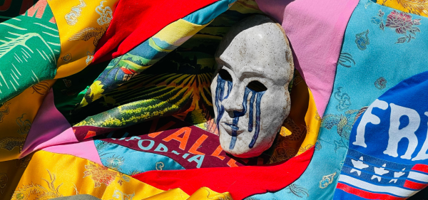 Theatrical grey mask with blue tears running down its cheeks on top of a multicolored quilt made of traditional Chinese fabrics.