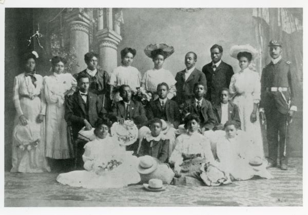 An old black-and-white photograph of 18 formally-dressed African American men and women standing and sitting in three rows with a painted theatrical background of Greco-Roman pillars