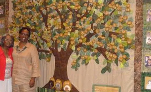 Two African American women, one with shoulder-length gray hair and one with short black hair, stand in front of a quilt portraying a tree with many golden fruits in its branches. 