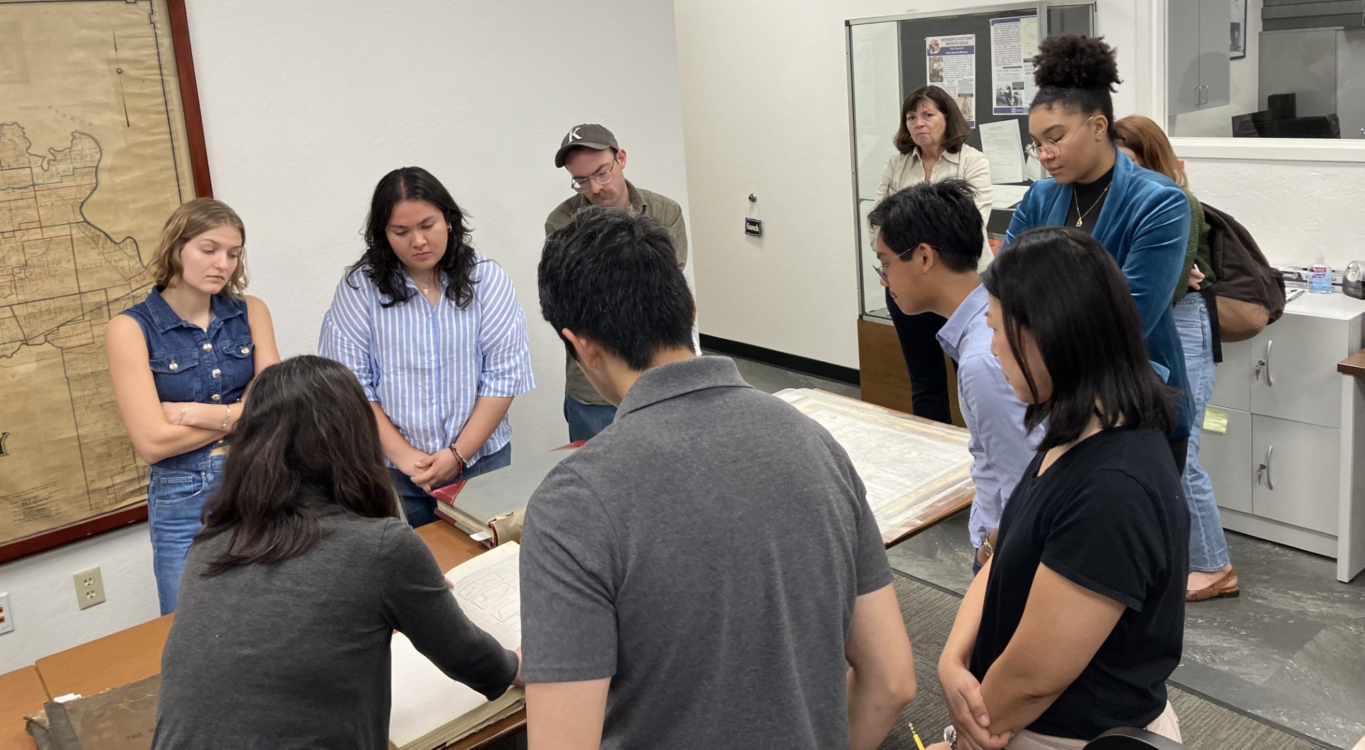 A group of 8 young adult UC Davis students standing around a table with historical documents at the Yolo County Archives, accompanied by an archivist pointing to one of the books. 