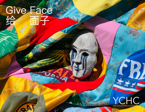 Give Face. Grey clay mask with blue tears streaming from the eye sockets down its cheeks propped up on a quilt made of traditional Chinese silk fabric and American tourist t-shirts.